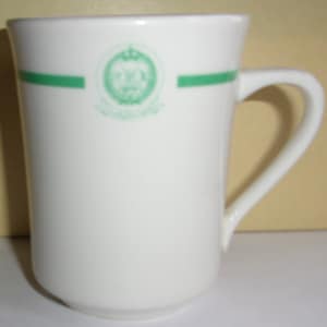 May include: White ceramic coffee mug with a green band and a green emblem featuring a crown and a sword. The emblem has Arabic text.