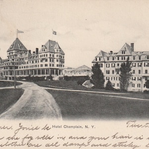 Op de afbeelding: Vintage zwart-wit ansichtkaart met het Hotel Champlain in New York. De afbeelding toont twee grote gebouwen met meerdere verdiepingen, vlaggen en een kronkelende weg die naar de ingang leidt. Handgeschreven tekst is zichtbaar onderaan.