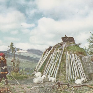 May include: Vintage postcard depicting a traditional dwelling with a grass roof and wooden supports. A person in traditional clothing stands near a structure with white goats resting nearby. Mountains and a cloudy sky form the backdrop.
