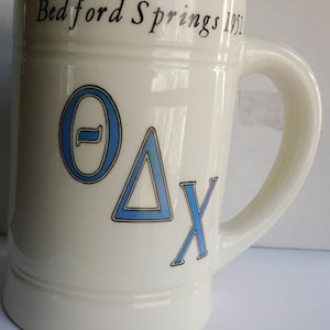 May include: White ceramic mug with blue and black lettering. The mug says "Bedford Springs 1952" and has the Greek letters "Theta Delta Chi" on the front.