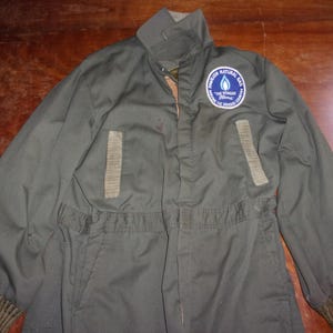 May include: Olive green jumpsuit with a zip-up front, collar, and ribbed cuffs. Features two rectangular patches and a circular patch with the text "Pioneer Natural Gas" and "The Wonder Flame" logo.