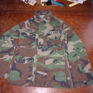 May include: Camouflage pattern military jacket with the name "TURNER" above the left pocket and "U.S. ARMY" above the right pocket. The jacket features multiple pockets and a button-down front. The color scheme includes shades of green, brown, and black.