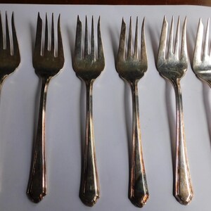 May include: Six silver forks with ornate handles. The forks are arranged in a row on a white surface.