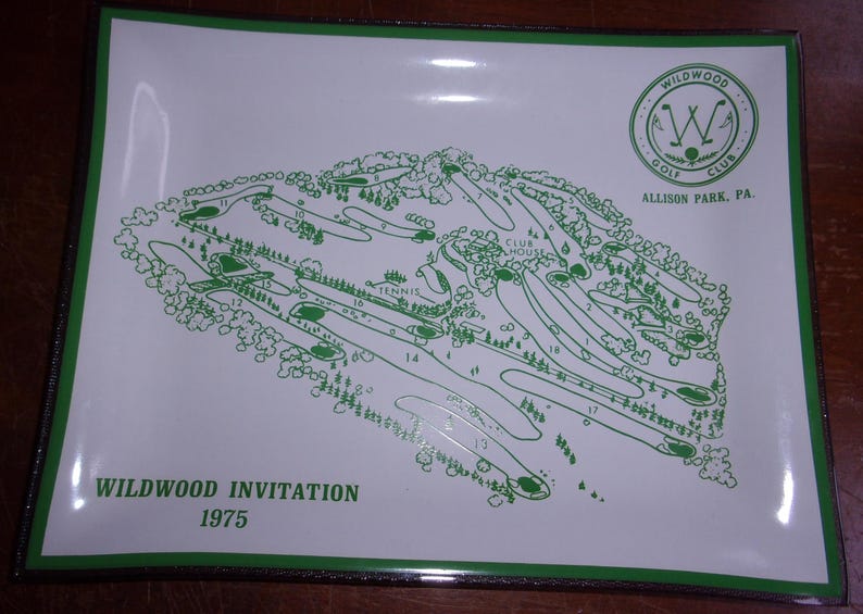 May include: A white rectangular tray with a green border displays a green golf course map. The map includes numbered holes, trees, and the text "Wildwood Invitation 1975." The Wildwood Golf Club logo is in the upper right corner.