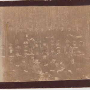 May include: A vintage sepia-toned photograph of a group of people, mostly children, standing in a wooded area. The group is arranged in rows, with the adults in the back row and the children in the front rows. The children are all wearing hats and some are holding small objects. The photograph is faded and has a warm, nostalgic feel.