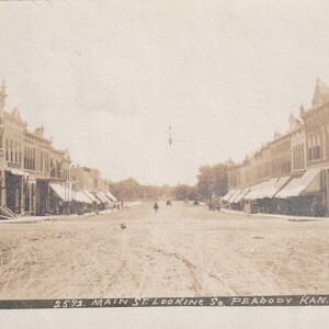 May include: Sepia-toned vintage postcard depicting Main Street in Peabody, Kansas. The image shows a wide dirt road flanked by two rows of multi-story buildings with awnings. The text at the bottom reads "2593. MAIN ST. LOOKING SE. PEABODY KAN."
