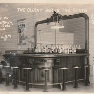 Peut inclure: Photographie en noir et blanc d'un bar vintage, le Loop Cafe à Georgetown, Colorado. Le bar comprend un grand miroir, des bouteilles et des tabourets. Le texte sur le mur indique "The Oldest Bar in the State" et "Brought to Georgetown, Colo in 1860 by Ox Team."
