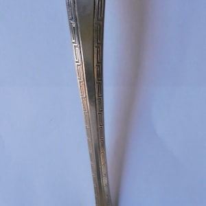 May include: A silver serving spoon with a square bowl and a long, ornate handle. The handle has a geometric pattern.