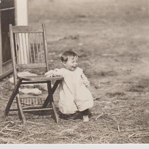 Puede incluir: Una fotografía en blanco y negro de un niño pequeño sentado en una silla plegable de madera. El niño lleva un vestido blanco y sonríe. La foto fue tomada al aire libre y el fondo está borroso.