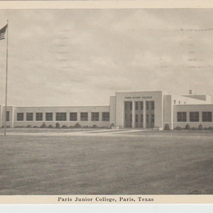 May include: A black and white photo of Paris Junior College in Paris, Texas. The building is a long, low structure with a flagpole in front. The flagpole is flying the American flag.