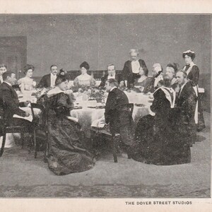 May include: A black and white photograph of a formal dinner party. A large round table is set with a white tablecloth and surrounded by people in formal attire. The Dover Street Studios is printed at the bottom.