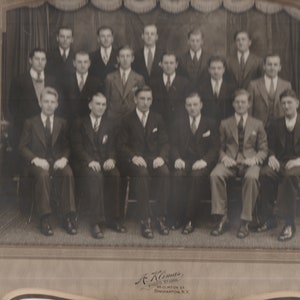 May include: Black and white photo of a group of men in suits, standing and sitting in front of a curtain. The men are all smiling and looking at the camera. The photo is framed and has a signature at the bottom.