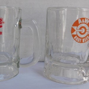 May include: Two glass mugs for root beer. The mug on the left has a red "Frostie Root Beer" logo. The mug on the right has an orange and white "A&W Root Beer" logo.