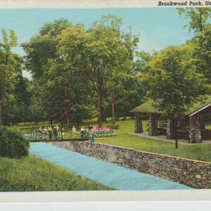 May include: A vintage postcard featuring a scenic view of Brookwood Park in Herkimer, New York. The postcard shows a lush green park with a stone bridge, a stream, and a stone gazebo. The park is surrounded by trees and foliage.