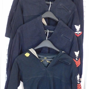 May include: Four navy blue sweaters with white collars and various insignia on the sleeves. The sweaters are hanging on hangers.
