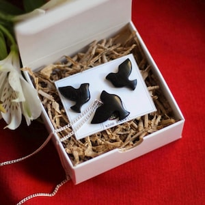 May include: A set of black bird-shaped earrings and a necklace in a white box with brown paper filler. The necklace is a silver chain with a black bird pendant.