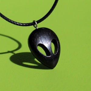 Può includere: Collana con ciondolo alieno nero con cordino. Il ciondolo presenta una testa di alieno scolpita con grandi aperture ovali per gli occhi. La collana è esposta su uno sfondo verde brillante.