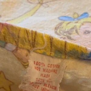 K&ouml;nnte beinhalten: Ein wei&szlig;es Stoff-Etikett mit dem Text "100% Baumwolle, hei&szlig; waschen, Schleudern, nicht bleichen, nicht b&uuml;geln" auf einem gelben und wei&szlig;en Stoffhintergrund mit einem Cartoon-Bild eines M&auml;dchens.