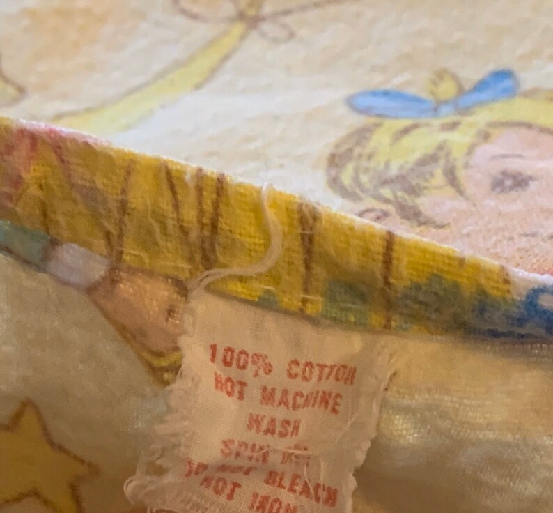 K&ouml;nnte beinhalten: Ein wei&szlig;es Stoff-Etikett mit dem Text "100% Baumwolle, hei&szlig; waschen, Schleudern, nicht bleichen, nicht b&uuml;geln" auf einem gelben und wei&szlig;en Stoffhintergrund mit einem Cartoon-Bild eines M&auml;dchens.