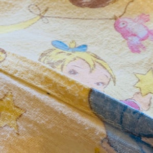 K&ouml;nnte beinhalten: Ein gelber Stoff mit einer Cartoon-Illustration eines M&auml;dchens mit blonden Haaren, das eine blaue Schleife tr&auml;gt. Das M&auml;dchen h&auml;lt einen Stern und ist von anderen Cartoon-Charakteren umgeben.