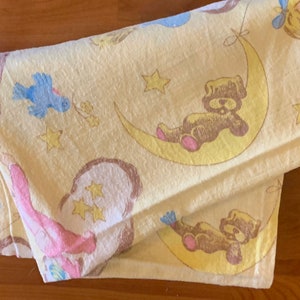 K&ouml;nnte beinhalten: Eine gelbe Babydecke mit einem braunen B&auml;ren, einem blauen Vogel und Sternen darauf gedruckt.