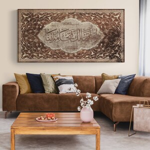 May include: A brown leather sectional sofa with a variety of throw pillows in different colors and patterns. A wooden coffee table with a vase of white cotton flowers sits in front of the sofa. A framed piece of art with Arabic calligraphy and intricate floral designs hangs on the wall behind the sofa.