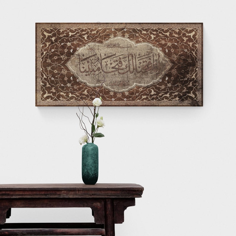 May include: A brown wooden framed canvas with intricate white floral designs and Arabic calligraphy in a white oval. The canvas is hanging on a white wall above a dark wooden table with a teal vase and white flowers.