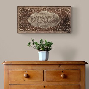 May include: A brown wooden framed artwork with intricate floral designs and Arabic calligraphy in a white oval. The artwork is hanging on a beige wall above a wooden dresser with drawers.
