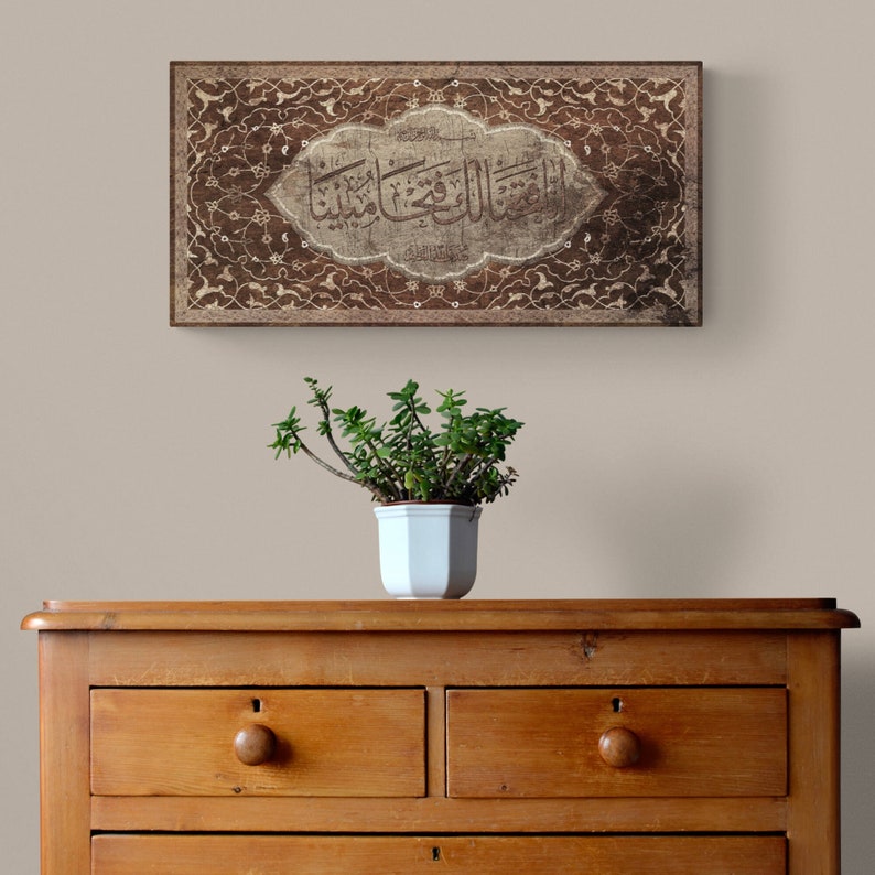 May include: A brown wooden framed artwork with intricate floral designs and Arabic calligraphy in a white oval. The artwork is hanging on a beige wall above a wooden dresser with drawers.