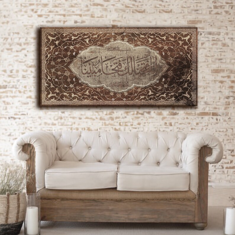 May include: A brown canvas print with a white background featuring Arabic calligraphy in a decorative frame. The frame is made of intricate floral designs in brown and gold. The print is hanging on a white brick wall above a white couch.