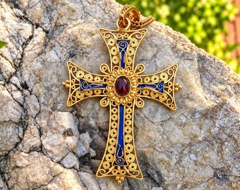 May include: A gold-toned metal cross pendant featuring intricate filigree scrollwork, vibrant blue enamel accents, and a central oval-shaped deep red gemstone. The cross has a decorative bail at the top and a detailed, ornate openwork design.