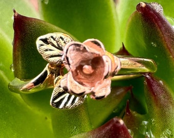 May include: A gold ring with a rose-colored flower design. The ring is set against a green succulent plant.