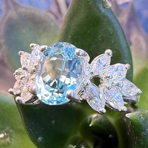 May include: A silver ring featuring a large oval blue topaz gemstone, surrounded by smaller clear gemstones. The ring is displayed against a green leafy backdrop.