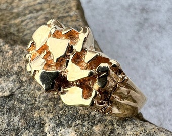May include: A gold ring with a textured, rough surface, resembling a rock formation. The ring is thick and has a wide band.