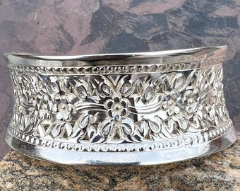 May include: A silver, decorative, floral patterned cake stand. The stand is round and has a raised edge.