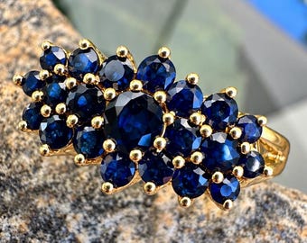 May include: A gold ring featuring a large central round-cut dark blue gemstone surrounded by a cluster of smaller round-cut dark blue gemstones. The stones are set in a gold band, creating a floral-like arrangement across the top surface.