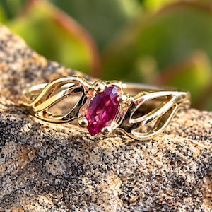 May include: A gold ring featuring a marquise-cut deep pink gemstone set in the center. The band is crafted with an open, swirling metal design that extends from the central stone, creating a delicate and intricate appearance.