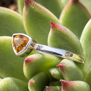 May include: A silver ring featuring a single triangular-cut amber-colored gemstone set in a smooth bezel. The band is polished silver with a reflective surface, showcasing a clean, metallic finish that contrasts with the warm, faceted orange stone.
