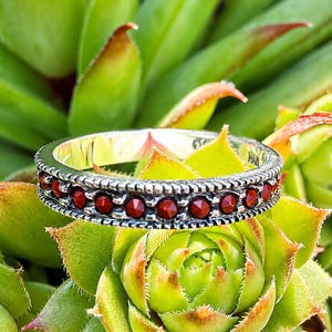 May include: A silver-toned metal ring featuring a continuous row of small, round, deep red faceted stones set within a textured, beaded border. The band has a metallic finish and a uniform width, showcasing a series of vibrant red accents.