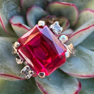 May include: A silver ring featuring a large, square, red gemstone and smaller, clear gemstones on the band.