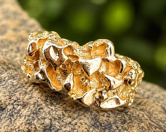 May include: A gold ring featuring a textured, nugget-style design with an irregular, bumpy surface. The metal has a polished, reflective finish that catches the light, creating deep shadows and bright highlights across the uneven, organic shape.