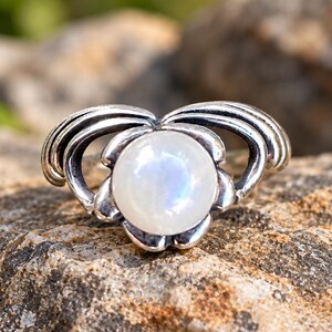 May include: A silver ring featuring a central, round, translucent white moonstone cabochon. The metal setting is shaped like stylized petals or wings that curve outward from the stone, creating a symmetrical, layered design with oxidized details.