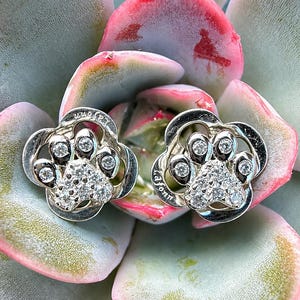 May include: A pair of silver-toned stud earrings shaped like paw prints, each featuring a central cluster of small clear stones and individual stones set into the toes. The metal surface is engraved with the brand name Lafonn on the side.