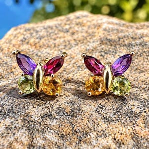 May include: A pair of gold butterfly-shaped stud earrings featuring marquise-cut purple and magenta gemstones for the upper wings and round-cut yellow and light green gemstones for the lower wings, set in a polished gold metal frame.