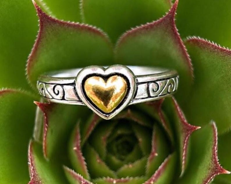 May include: A silver-toned ring featuring a central heart-shaped element with a polished gold-toned finish. The band is crafted with an oxidized silver-toned metal and displays a repeating embossed scrollwork pattern along the sides of the ring.