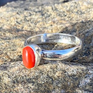 Pu&ograve; includere: Un anello d'argento con una pietra preziosa ovale di colore rosso brillante. L'anello &egrave; posto su una superficie rocciosa.