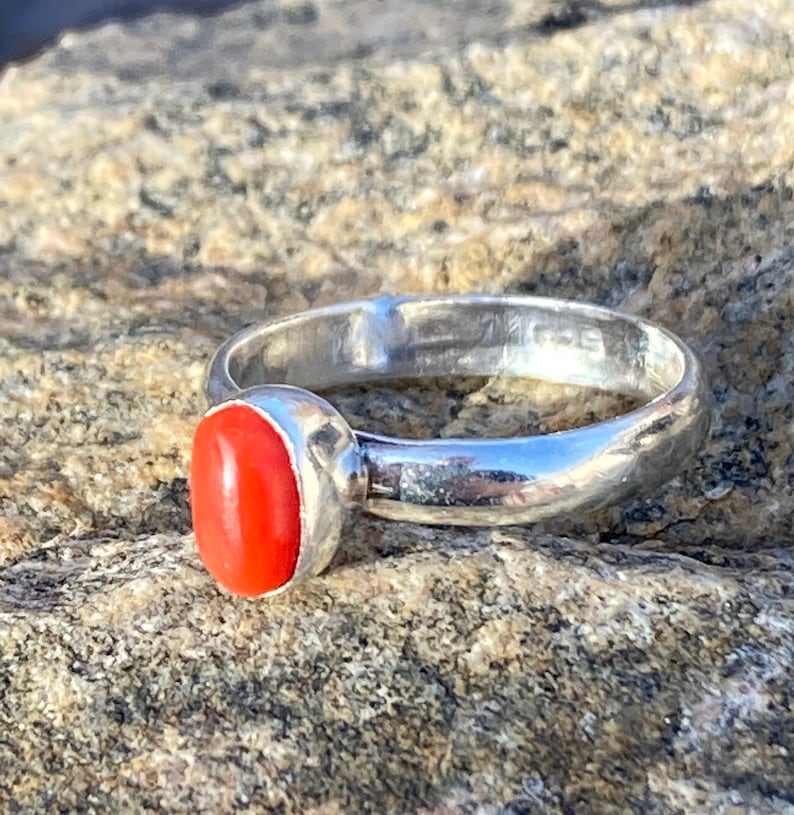 Pu&ograve; includere: Un anello d'argento con una pietra preziosa ovale di colore rosso brillante. L'anello &egrave; posto su una superficie rocciosa.