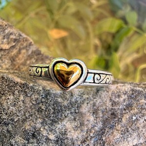 May include: A silver-toned ring featuring a central polished gold-toned heart element. The band is crafted with a dark oxidized silver finish and displays a repeating embossed scrollwork pattern that extends across the visible portion of the ring.