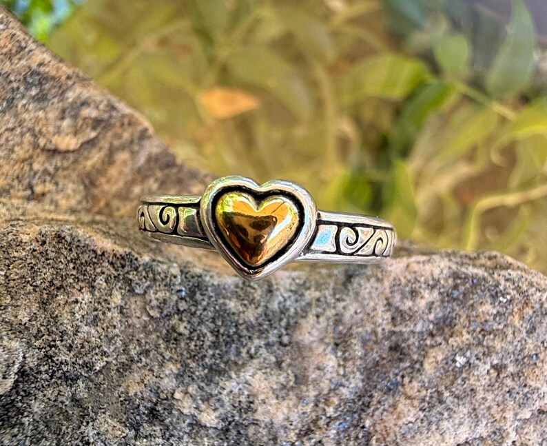May include: A silver-toned ring featuring a central polished gold-toned heart element. The band is crafted with a dark oxidized silver finish and displays a repeating embossed scrollwork pattern that extends across the visible portion of the ring.