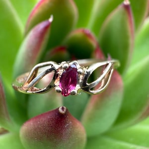 May include: A gold ring featuring a marquise-cut, deep pink gemstone. The ring has an openwork design on either side of the gemstone. The ring is set against a backdrop of green and red succulent leaves.